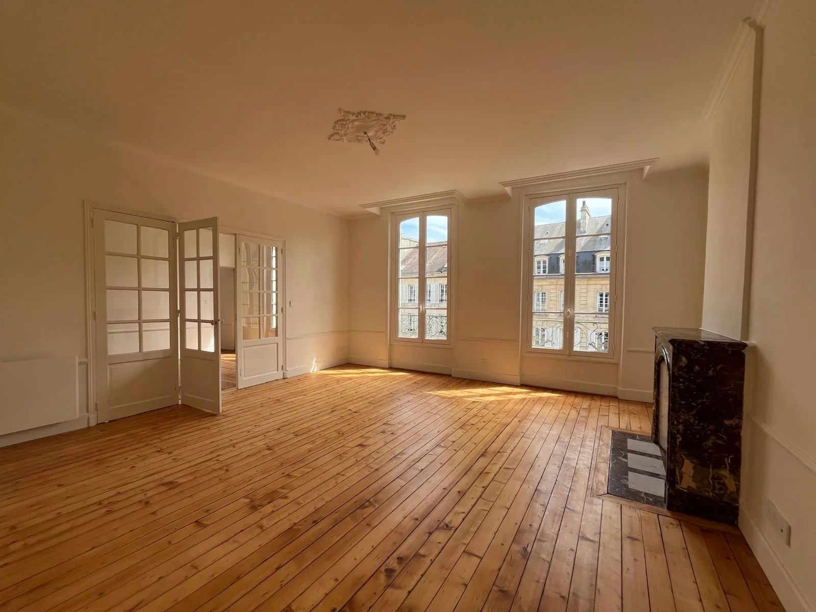 Grande pièce avec rosace, corniches et moulures murales claires, parquet bois et cheminée en marbre.
